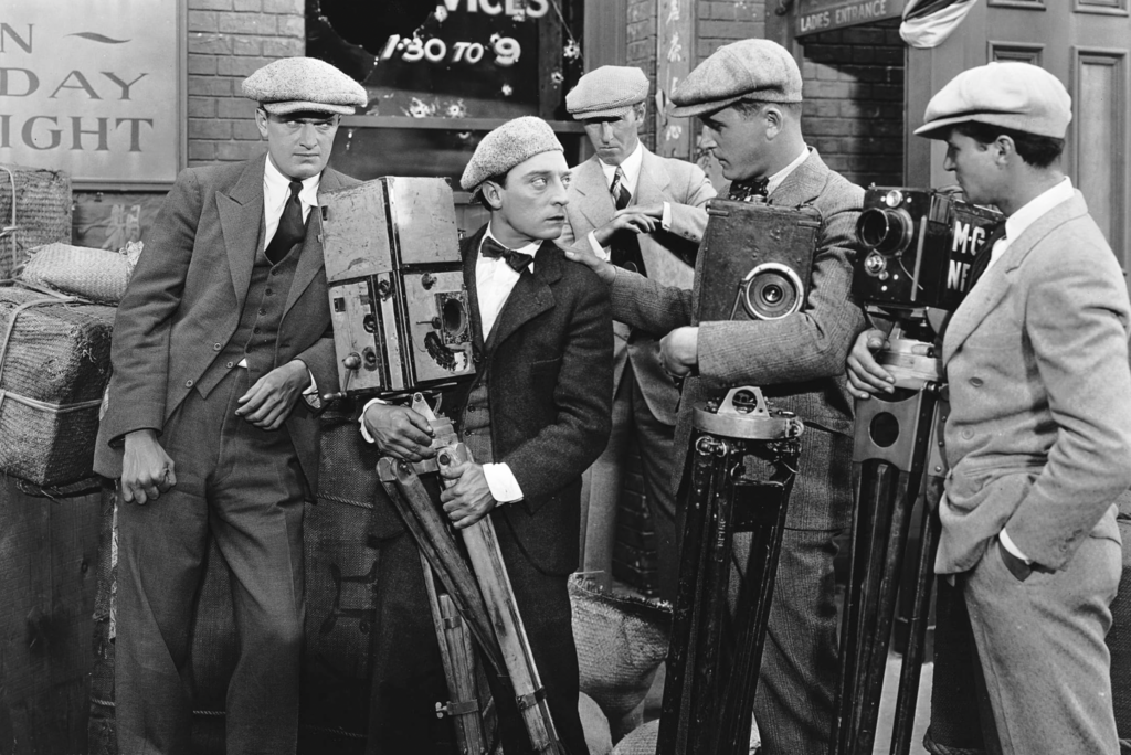 Black and white still from "The Cameraman" featuring Buster Keaton holding a camera and surrounded by other cameramen.