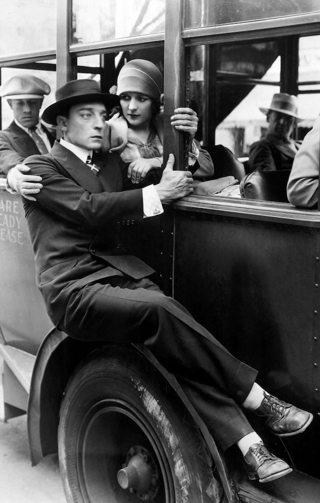 Black and white still from "The Cameraman" featuring Buster Keaton sitting on the exterior of a bus to be near Marcelline Day's character.