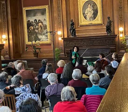 A group of people listen to a musical performance in the Robbins Library Reading Room.